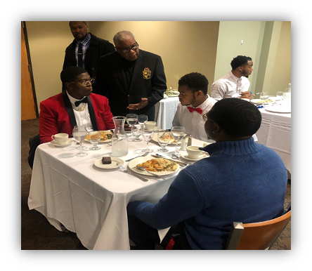 Three people sitting at dinner table being talked to by a standing man.