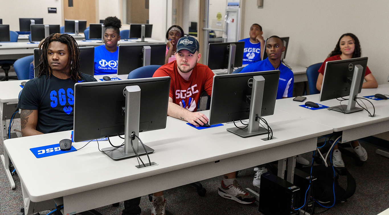 A group of students sit in front of computers in a computer lab while attending class