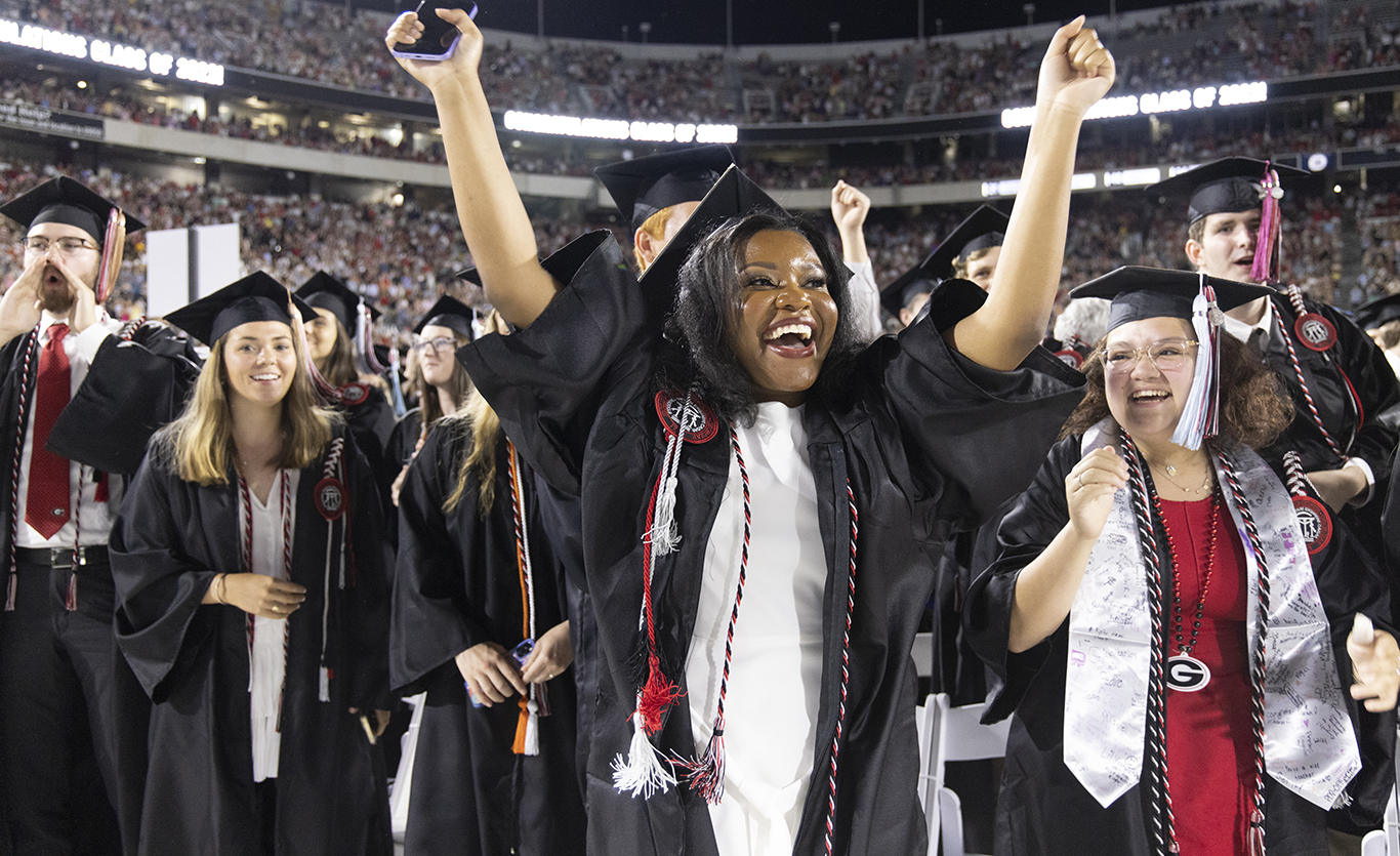 A grinning young woman in cap and gown raises her arms in celebration amongst a group of fellow graduates at graduation
