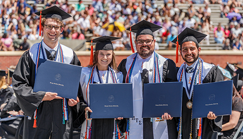 Four graduates hold up their diplomas at graduation
