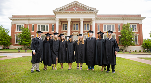 Nine students in graduate regalia stand in front of a university building