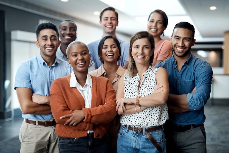 A diverse group of people with arms crossed, wearing business casual in an office setting and smiling for the camera