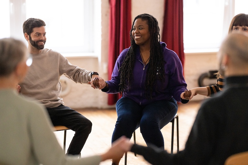 Five people seated in chairs sit in a circle holding hands during a group session