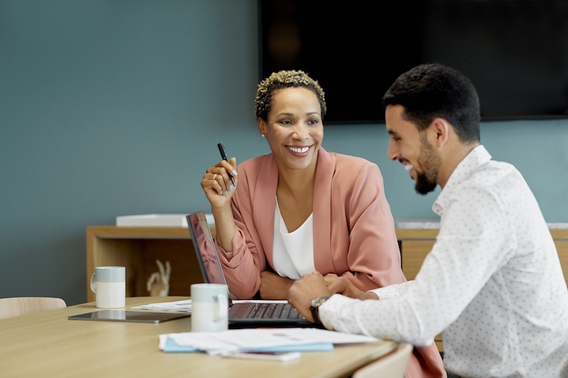 A smiling man sits in front of a laptop at a conference table next to a smiling woman with a pen and papers