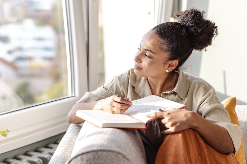 A relaxed woman sitting by a window while writing in a notebook