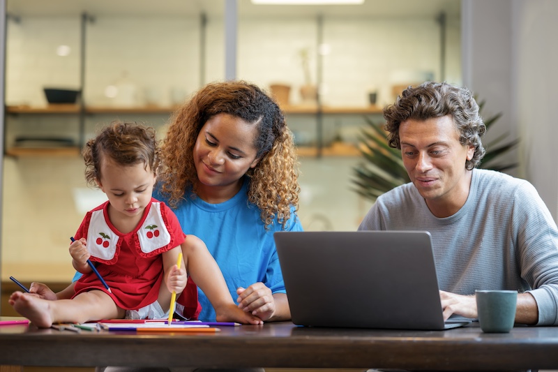 A couple sits at a dining table while they work on a laptop and take care of a small child